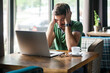© khosrork - Portrait of exhausted tired man freelancer wearing green T-shirt, suffering migraine or high blood pressure while working online on laptop, Indoor shot near big window, cafe background.