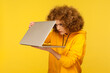 Curious woman with Afro hairstyle peeking into half-opened laptop and expressing amazement, spying on forbidden content, wearing casual style hoodie. Indoor studio shot isolated on yellow background.