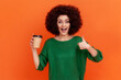 © khosrork - Excited woman with Afro hairstyle wearing green casual style sweater holding coffee to go and showing thumb up, recommend coffee house. Indoor studio shot isolated on orange background.
