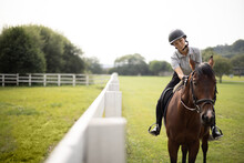 Female Horseman Riding Brown Thoroughbred Horse On Green Meadow Near Fence In Countryside. Concept Of Rural Resting And Leisure. Idea Of Green Tourism. Young Smiling European Woman. Sunny Daytime