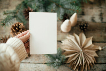 hand holding empty greeting card on background of christmas paper stars, wooden tree, pine branches 