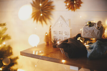 Wall Mural - Stylish zero waste christmas gifts on wooden shelf on background of little house, trees, paper stars and christmas golden lights bokeh. Festive decorated scandinavian room. Atmospheric eve