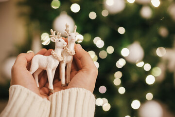 Wall Mural - Hands holding stylish reindeer toy on background of christmas tree lights. Beautiful christmas deer toy in hands in festive decorated scandinavian room. Space for text. Magic winter holidays