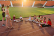 A large group of children, boys and girls, sit and listen to the coach's instructions before the game at the stadium during sunset. A healthy lifestyle.