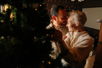 Wall Mural - Loving young couple decorating Christmas tree with shiny balls standing hugging near fireplace in cozy dark living room. Happy man and woman preparing home for New Year holidays celebration