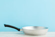 New aluminium frying pan on wooden table at light blue wall background. Pastel color. Closeup. Cooking concept. Empty place for text. Front view.