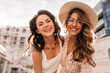 © Look! - Close-up photo of two young fair-skinned girls taking photo against blurred sky background. Fair-haired lady with transparent glasses and hat on her head. Brunette in white top with red necklace.