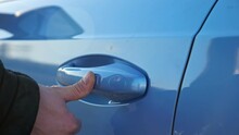 Driver in warm black jacket uses keyless entry access by putting finger on handle to open or close blue foreign car with fingerprint closeup
