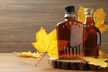 bottles of tasty maple syrup and dry leaves on wooden table, space for text