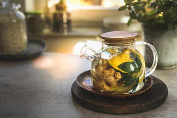 Glass tea pot with mint leaves, lemon and ginger on kitchen table with utensils and window background. Herbal tea with healthy ingredients and vitamin c in cold season. Natural treatment. Front view.