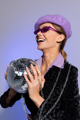 Wall Mural - trendy woman in beret, sunglasses and blazer with animal print holding disco ball and laughing isolated on violet