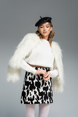 Wall Mural - young woman in beret, white faux fur jacket and skirt with animal print posing isolated on grey