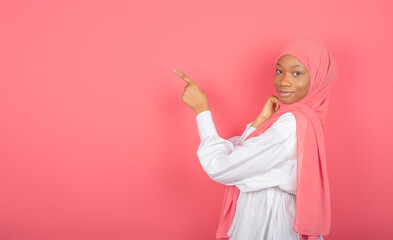 Young African American Muslim woman points index finger to free space wrapped in pink veil isolated on pink background.
