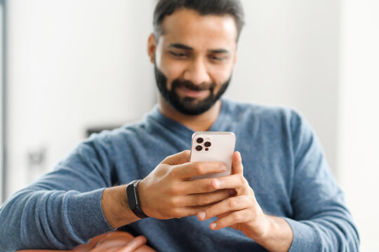 Selective focus at trendy smartphone in hands of concentrated young indian guy, hispanic male freelancer using phone for remote work from home, ethnic man using new mobile app, texting online