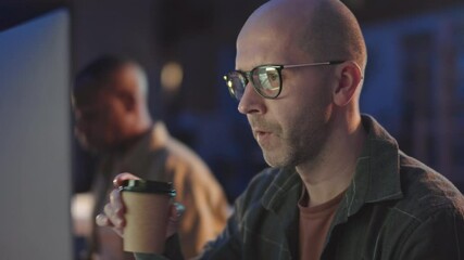 Medium close-up with slowmo of bald mid-adult programmer in eyeglasses working on computer late in dark office together with colleague