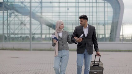 Wall Mural - Portrait of happy muslim couple, woman in hijab and manin jacket, walking with passports and tickets outdoors carrying a suitcase and talking each other, going to travel by airplane at modern airport.