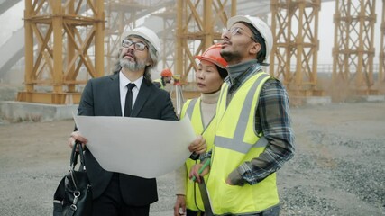 Wall Mural - Architect mature man in suit is discussing project with builders wearing uniform showing blueprint standing at construction site. People and job concept.