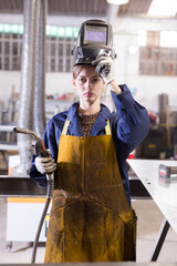 Young workwoman skilled welder wearing protective apron, gloves and helmet standing in metalworking workshop..