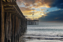 A Long Brown Wooden Pier With American Flags Flying On Curved Light Posts With People Walking And Fishing On The Pier With Powerful Clouds At Sunset At Ventura Pier In Ventura California USA