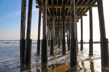 a shot of the legs underneath a brown wooden pier at the beach with waves rolling in from the vast blue ocean water with silky brown sand at Ventura Pier in Ventura California USA