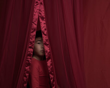 Studio Portrait Of Asian Girl In Red Dress Peeking Through Curtain Showing Part Of Her Face