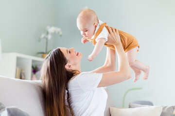 Wall Mural - Profile side view portrait of attractive careful cheerful girl playing with baby sitting on divan at light home flat house indoors