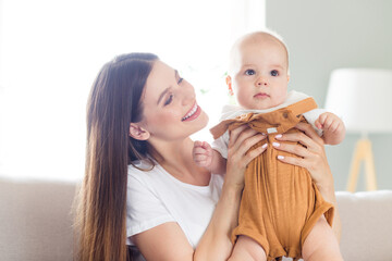 Sticker - Portrait of attractive careful cheerful girl carrying baby sitting on divan playing at light white home flat house indoors