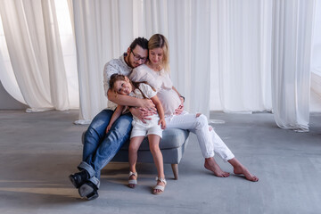 Wall Mural - In minimalist studio, pregnant young woman sits on a pouf, a bearded man hugs a little girl. Happy family in anticipation of a child. The father hugs the mother, the daughter laughs. Healthy pregnancy