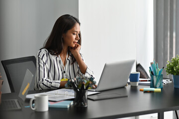 Photo of a young business owner looking at the computer laptop at her modern working desk surrounded by paperwork and office equipment.