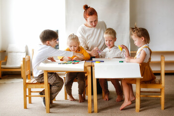 cheerful teacher woman with a group of cute preschool children at the table draw on paper with multi