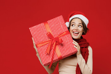 Young smiling fun woman 20s in Santa Claus Christmas red hat hold present box with gift ribbon bow look aside isolated on plain red background studio portrait. Happy New Year 2022 celebration concept.