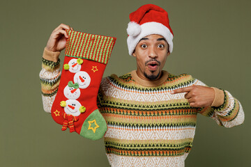 Young happy african man in knitted sweater red Christmas Santa Claus hat point on stocking gift sock isolated on plain green khaki background studio portrait. Happy New Year 2022 celebration concept.