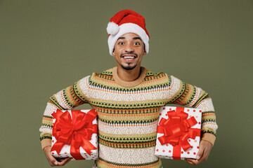 Young cheerful african man in sweater red Christmas Santa Claus hat hold red present box with gift ribbon bow isolated on plain green khaki background studio. Happy New Year 2022 celebration concept.