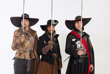 Cropped Portrait Of Thee Men In Vitage Costumes With Swords, Musketeer And Pirate Isolated Over White Background