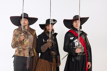 Wall Mural - Cropped portrait of thee men in vitage costumes with swords, musketeer and pirate isolated over white background