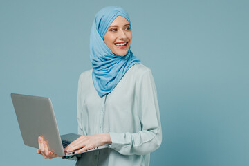 Wall Mural - Young arabian asian muslim woman in abaya hijab hold use work on laptop pc computer look aside on workspace isolated on plain blue background studio. People uae middle eastern islam religious concept.