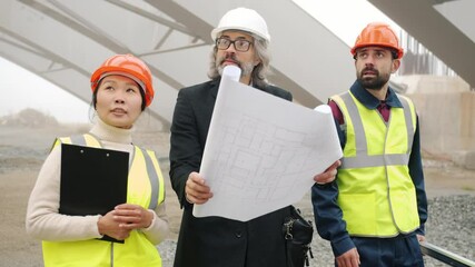 Wall Mural - Architect is talking to construction workers showing blueprint walking in building area outdoors. People are wearing safety uniform and helmets.