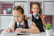 © dvulikaia - Lifestyle school portrait. Schoolgirl doing task sitting at desk, another girl doing bunny ears for her.