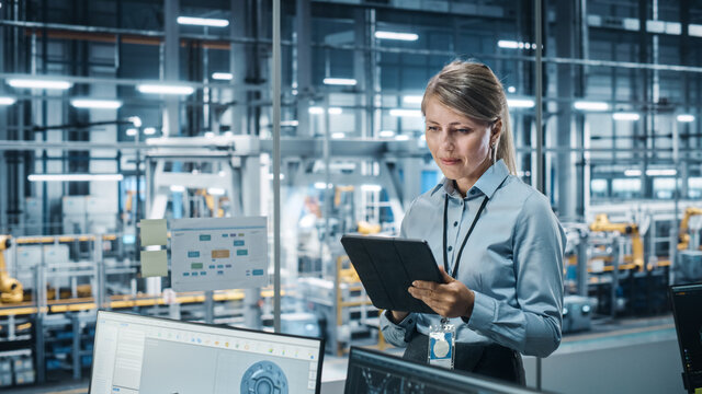Car Factory Office Concept: Portrait of Successful Female Chief Automotive Engineer Using Tablet Computer to Design and Optimize Automated Robot Arm Assembly Line Manufacturing Electric Vehicles