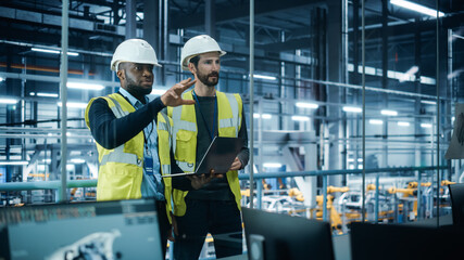 Leinwandbilder - Car Factory: Project Manager and Automotive Engineer in Hard Hat, Talking, Using Laptop. Monitoring, Control, Equipment Production. Automated Robot Arm Assembly Line Manufacturing Electric Vehicles