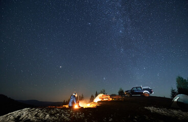 Wall Mural - Lonely woman sitting on grass by burning fire near two tents and black SUV. Night camping on mountain hill under starfull sky. Tree tops and silhouettes of mountains afar on the background.