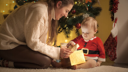 Happy mother giving Christmas present in golden box to her little baby son. Families and children celebrating winter holidays.