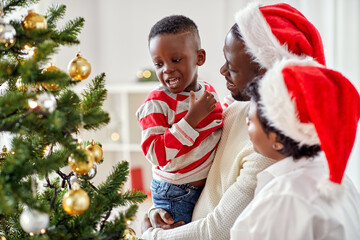 Wall Mural - family, winter holidays and people concept - happy african american mother, father and little son decorating christmas tree at home on