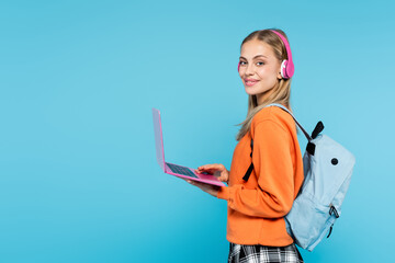 Wall Mural - Blonde student in headphones and backpack holding laptop isolated on blue