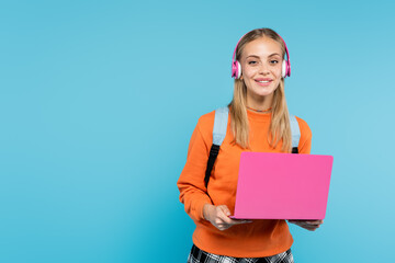Wall Mural - Smiling student in headphones holding laptop and looking at camera isolated on blue