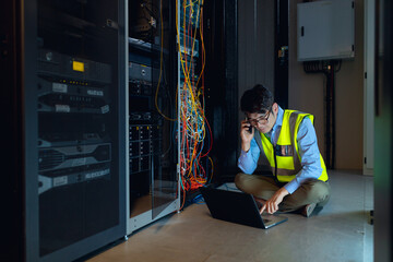 Asian male engineer using laptop and talking on smartphone while inspecting in computer server room