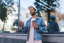 Happy Spanish Hipster Guy In Bluetooth Headphones Enjoying Positive Music Playlist From Mobile Radio Using 4g Wireless In City, Cheerful Male Tourist In Earbuds Holding Coffee To Go And Smartphone