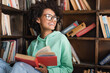 © LIGHTFIELD STUDIOS - happy african american student in eyeglasses sitting with book in library