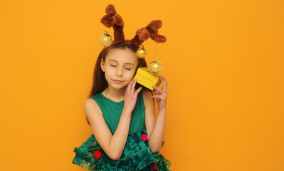 Girl with a deer ears in green festive dress holding a gift on yellow background with copy space. Christmas concept.