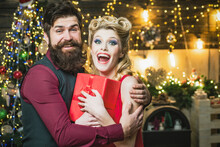Beautiful Christmas Couple With Gift Hugging. Retro Woman With Blonde Hair And Vintage Bearded Man Near The Christmas Tree.